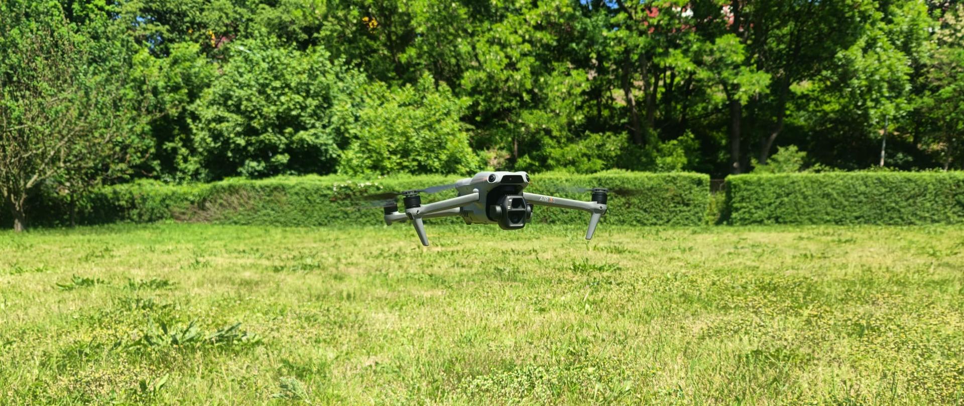otografia przedstawia bezzałogowy statek powietrzny (dron) marki DJI, model Air 3S, sfotografowany od przodu podczas zawisu w powietrzu. Dron znajduje się około metra nad ziemią, bezpośrednio nad rozłożonym na zielonej trawie ośmiokątnym, czarnym lądowiskiem marki Sunnylife z żółtym oznaczeniem lądowania „H”. Tło zdjęcia to jasny, nasłoneczniony trawnik, za którym ciągnie się ściana gęstej, zielonej roślinności, krzewów i drzew. Światło jest dzienne, mocne, charakterystyczne dla bezchmurnego, letniego dnia.