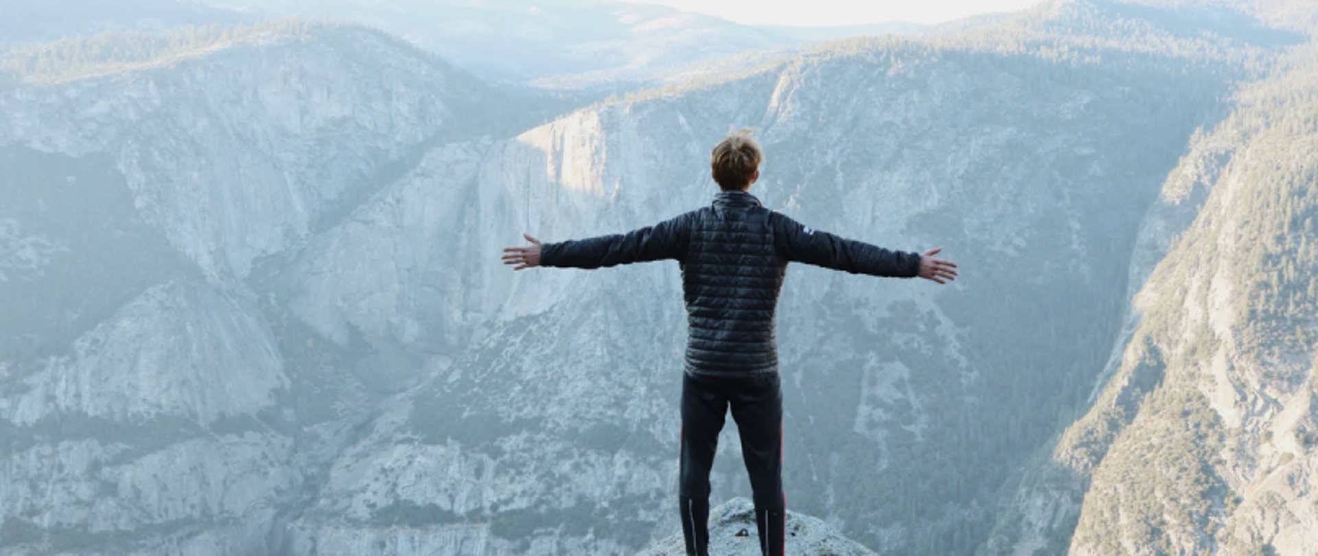 Człowiek na klifie z rozpostartymi ramionami, w tle góry i drzewa.
