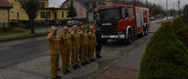Na zdjęciu widać strażaków ubranych w mundury koloru piaskowego. Strażacy stoją na zbiórce w szeregu. Jeden z nich salutuje. Po prawej stronie stoi samochód strażacki. Na dalszym planie widać ulicę oraz budynki mieszkalne
