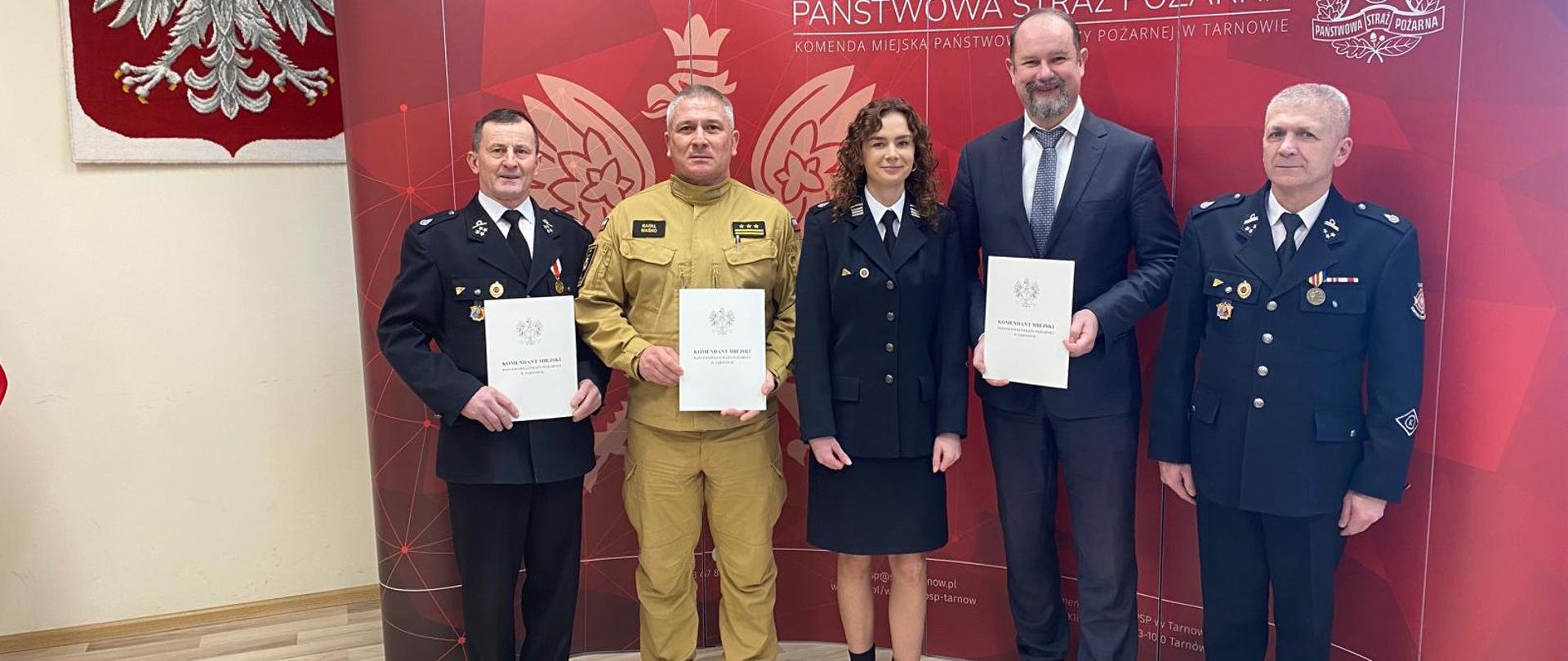 Przed czerwonym banerem Pastowej Straży Pożarnej stoją w rzędzie strażacy ubrani w mundury oraz wójt ubrany w garnitur. W rękach trzymają porozumienie.