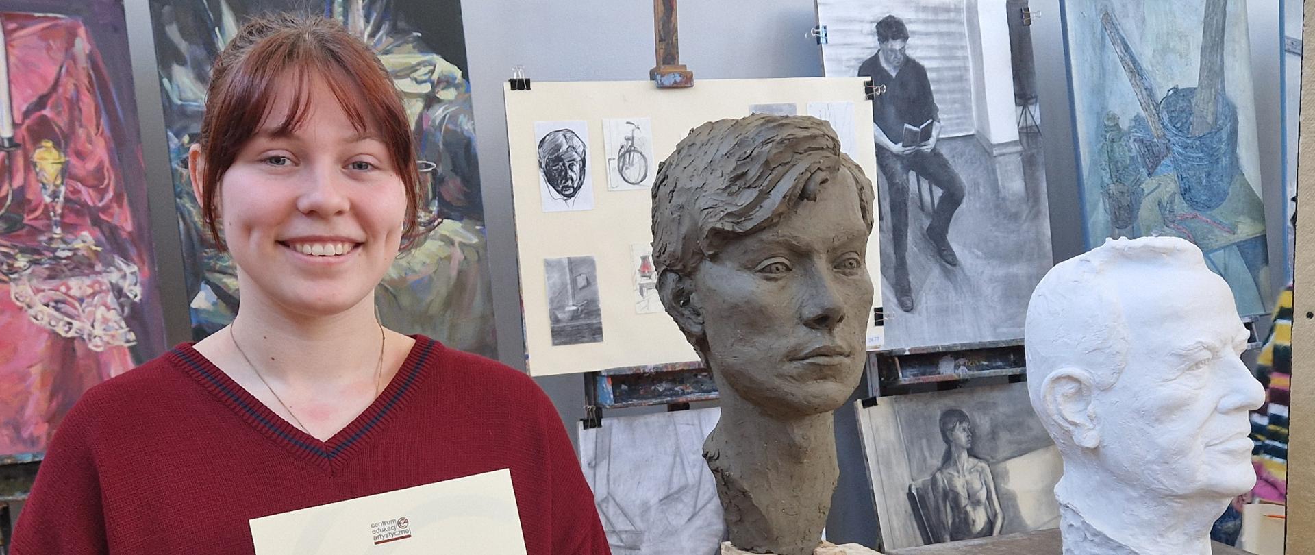 Na zdjęciu po lewej stronie widzimy Zuzannę Rumian uczennicę kl. 4 PLSP im. A. Kenara w Zakopanem. Zuzanna ubrana jest w brązowy sweter z niebieskim pasem na piersiach. Uczennica jest uśmiechnięta, trzyma w rękach dyplom za udział w drugim etapie Ogólnopolskiego Konkursu Plastycznego organizowanego przez Centrum Edukacji Artystycznej. Na dyplomie widnieją podpisy Jurorów. Obok Zuzanny widzimy dwie głowy zrealizowane przez uczennicę. Głowa pierwsza po lewej jest wykonana z gliny i przedstawia młodego mężczyznę. Druga głowa po prawej stronie wykonana jest z białego gipsu - to portret Stanisława Marduły wieloletniego nauczyciela lutnictwa w PLSP im. A. Kenara. Głowy stoją na stojakach. Z tyłu na ścianie widzimy prace rysunkowo-malarskie innego ucznia naszej szkoły Sebastiana Zycha biorącego udział w konkursie. 