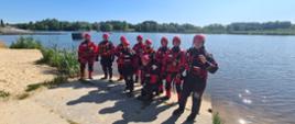 Strażacy biorący udział w szkoleniu ubrani w czerwone, specjalne skafandry do wody i kaski. Stoją nad brzegiem jeziora. 