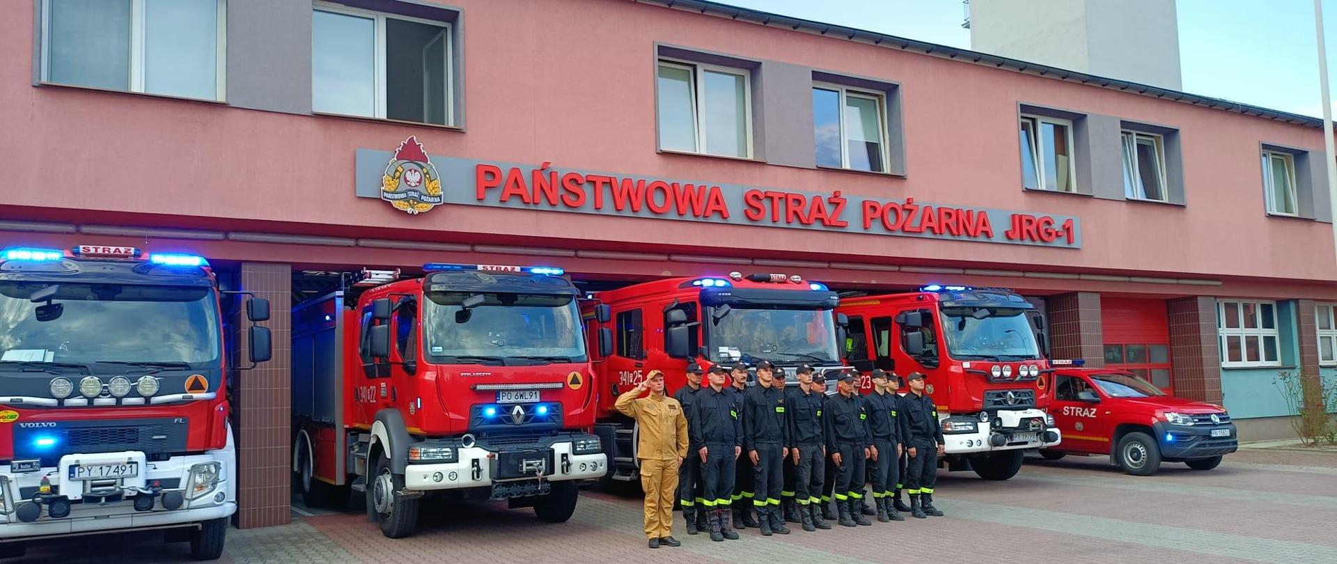 Strażacy w dwuszeregu stoją na tle samochodów strażackich