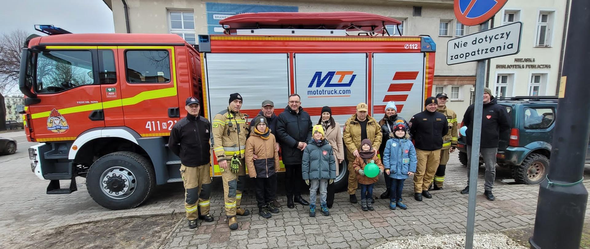 Przy dużym samochodzie strażackim, stoją strażacy i dzieci.