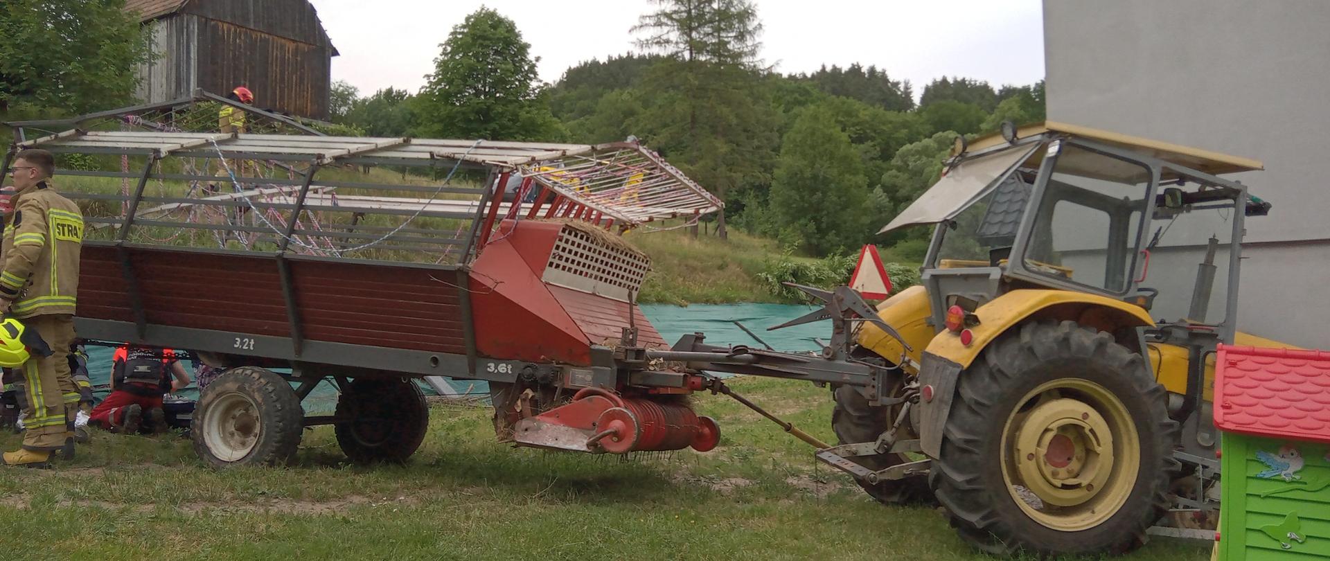 Zdjecie przedstawia ciągnik rolniczy wraz z przyczepą. Na zdjęciu znajdują się równie strażacy podczas akcji ratowniczej
