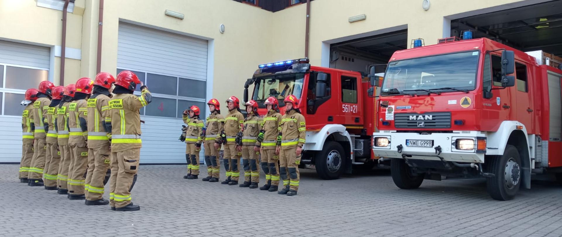 Strażacy przed budynkiem KP stoją naprzeciw siebie i oddają honor. Za nimi stoją pojazdy gaśnicze z włacoznymi sygnałami.