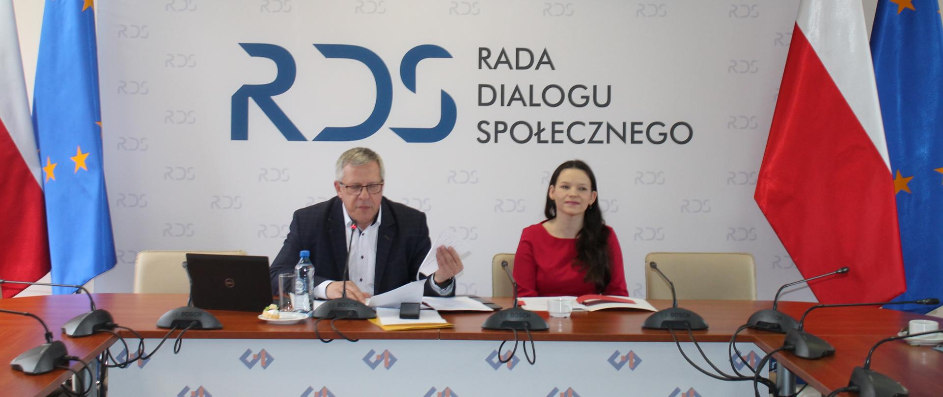 Na zdjęciu widnieje stół prezydialny za którym siedzą od lewej strony: Przewodniczący zespołu ds. budżetu, wynagrodzeń i świadczeń socjalnych oraz Katarzyna Nowakowska - Podsekretarz Stanu w Ministerstwie Rodziny, Pracy i Polityki Społecznej. Po obu stronach stołu widnieją dwie flagi: Polski i UE. W tle siedzących znajduje się baner z napisem Rada Dialogu Społecznego a nad nim wisi godło Polski.