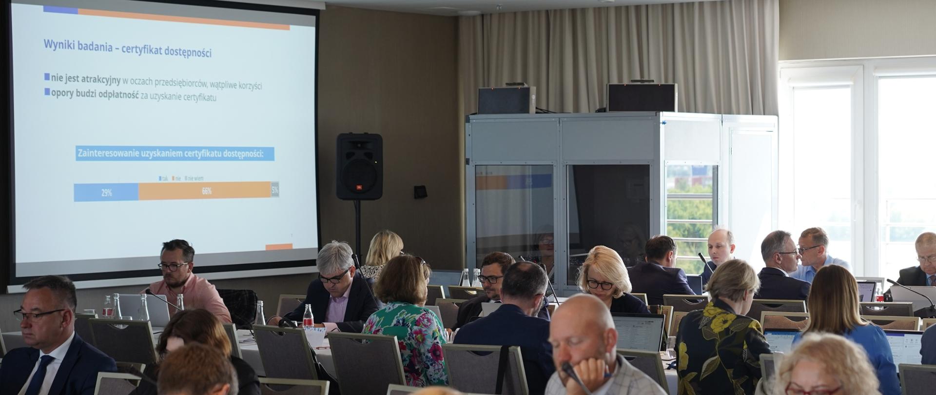 Grupa osób siedzi przy stołach na sali. 