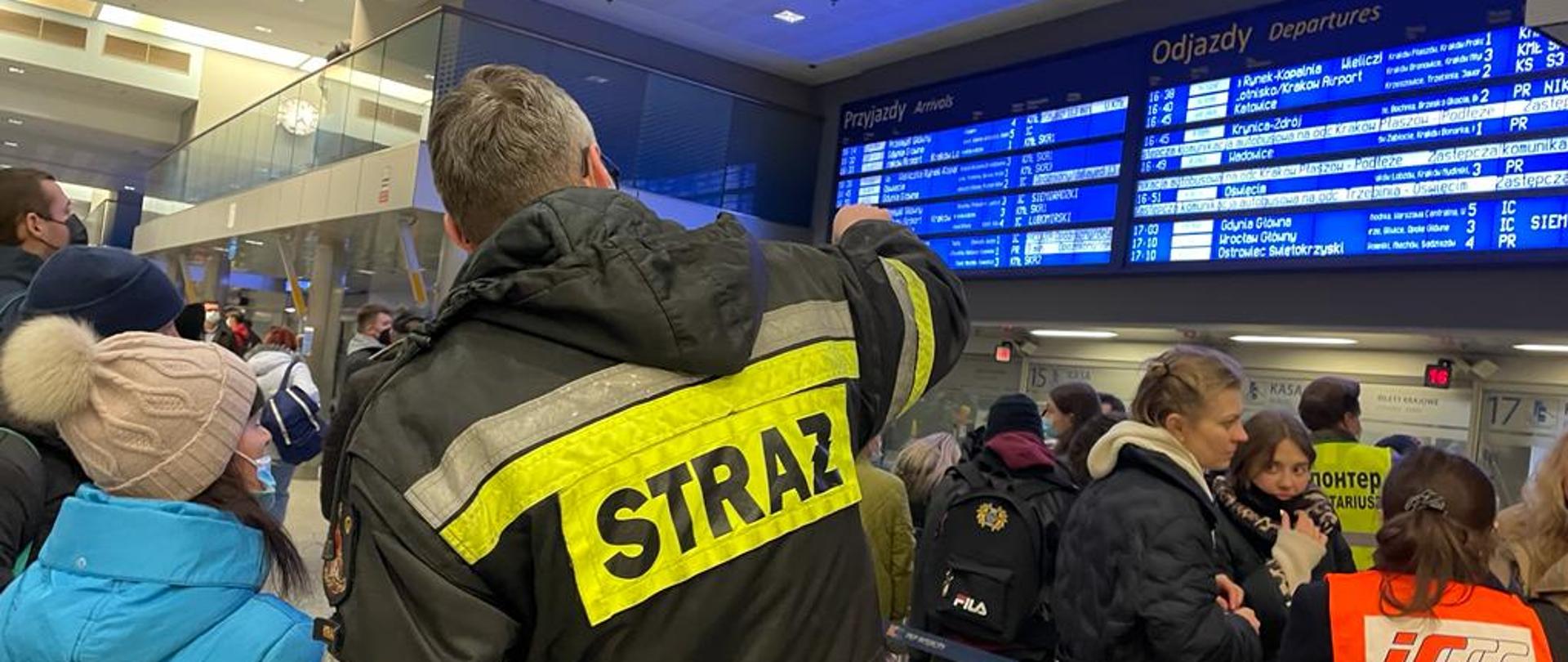 Strażak wskazuje kobiecie w niebieskiej kurtce godzinę odjazdu pociągu