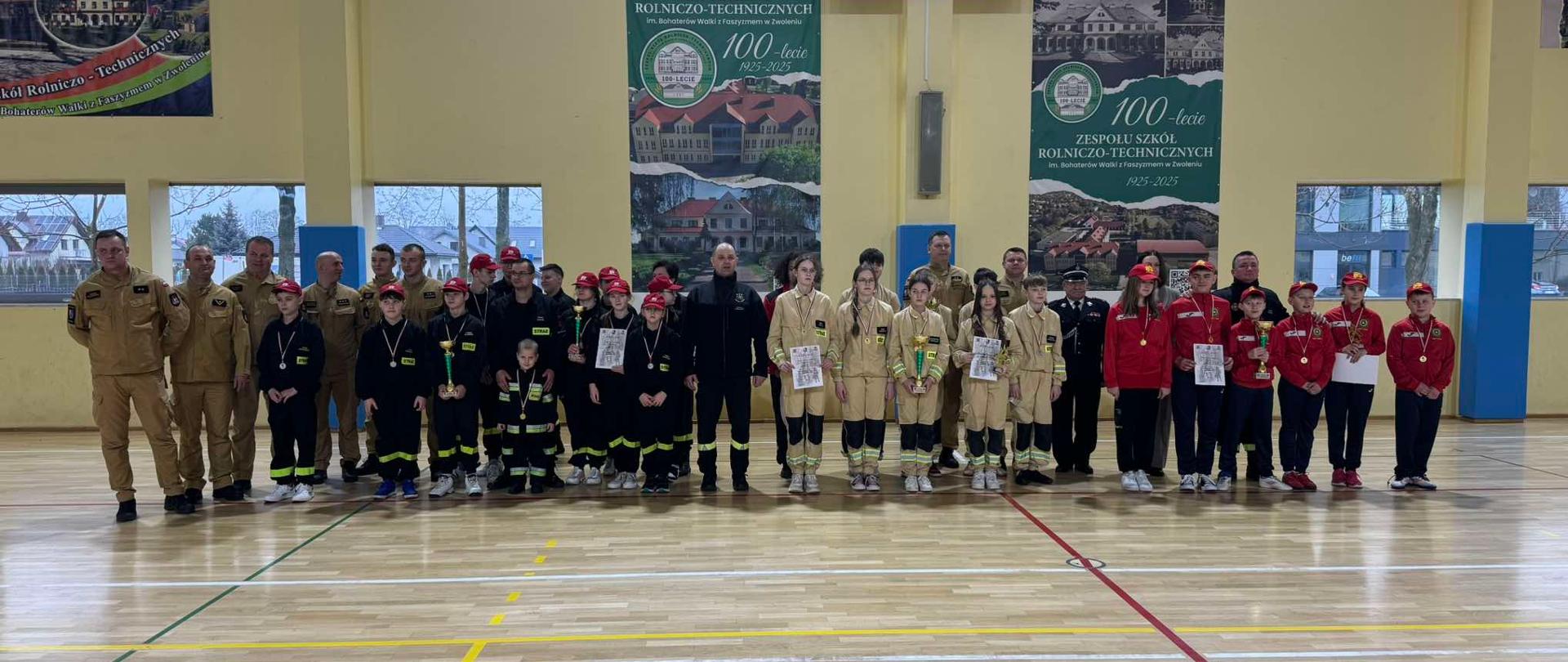 Zdjęcie przedstawia dużą grupę młodzieży i dorosłych, w tym strażaków w mundurach, zebranych na sali gimnastycznej. Młodzież ubrana jest w stroje sportowe i strażackie, niektórzy mają medale na szyjach i trzymają puchary, co sugeruje zawody lub uroczystość zakończenia szkolenia. W tle widoczne są banery upamiętniające 100-lecie Zespołu Szkół Rolniczo-Technicznych (1925-2025). Na ścianie wisi także tablica wyników.