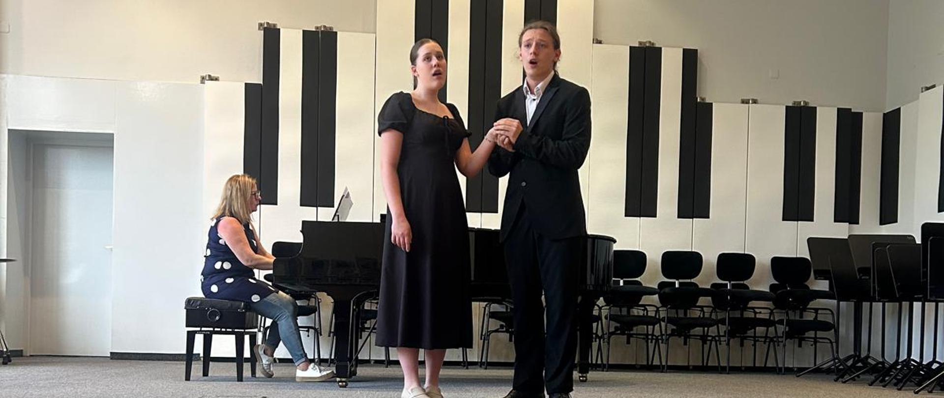 Dziewczyna i chłopak śpiewają, kobieta akompaniuje na fortepianie.