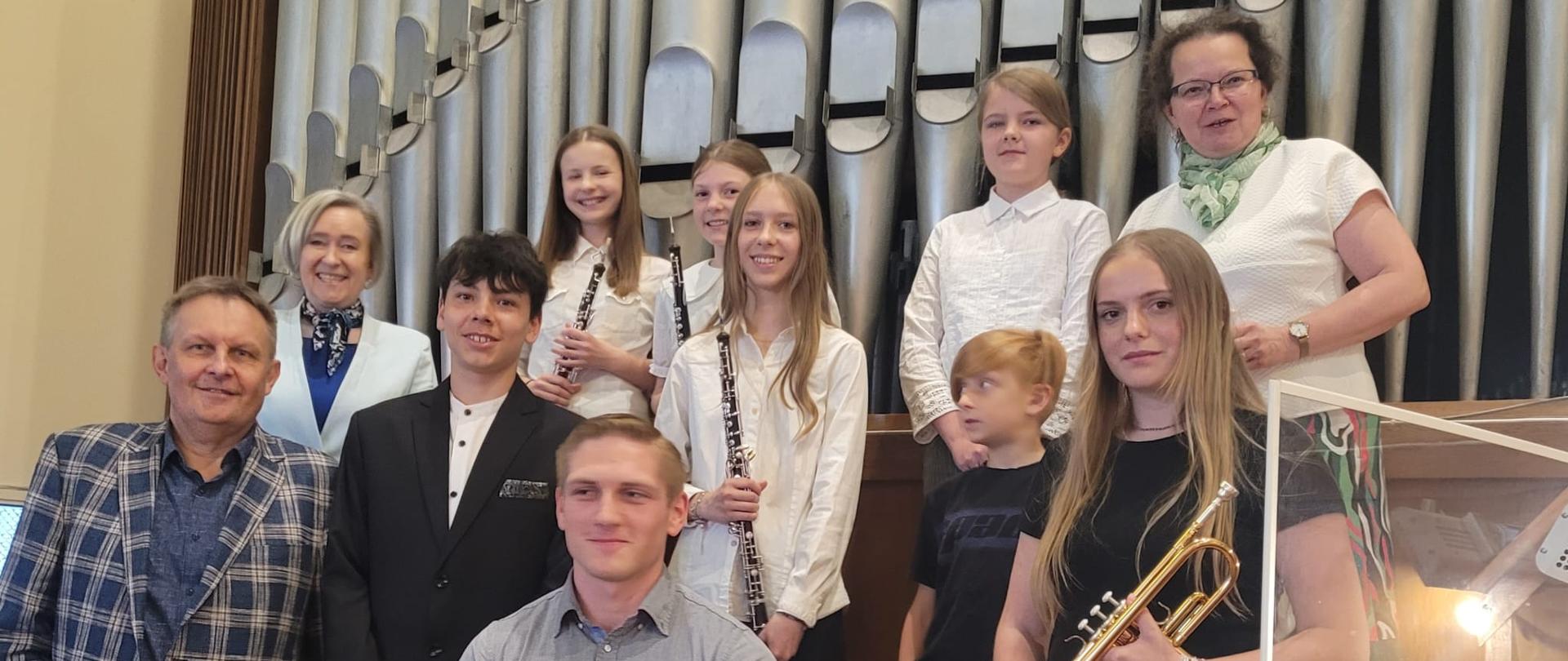 Pamiątkowe zdjęcie uczestników koncertu organowego w kościele parafialnym Najświętszego Serca Pana Jezusa w Gdyni. Na fotografii uczniowie szkół muzycznych wraz z pedagogami pozują na tle prospektu organowego; część uczestników trzyma instrumenty – oboje i trąbkę.