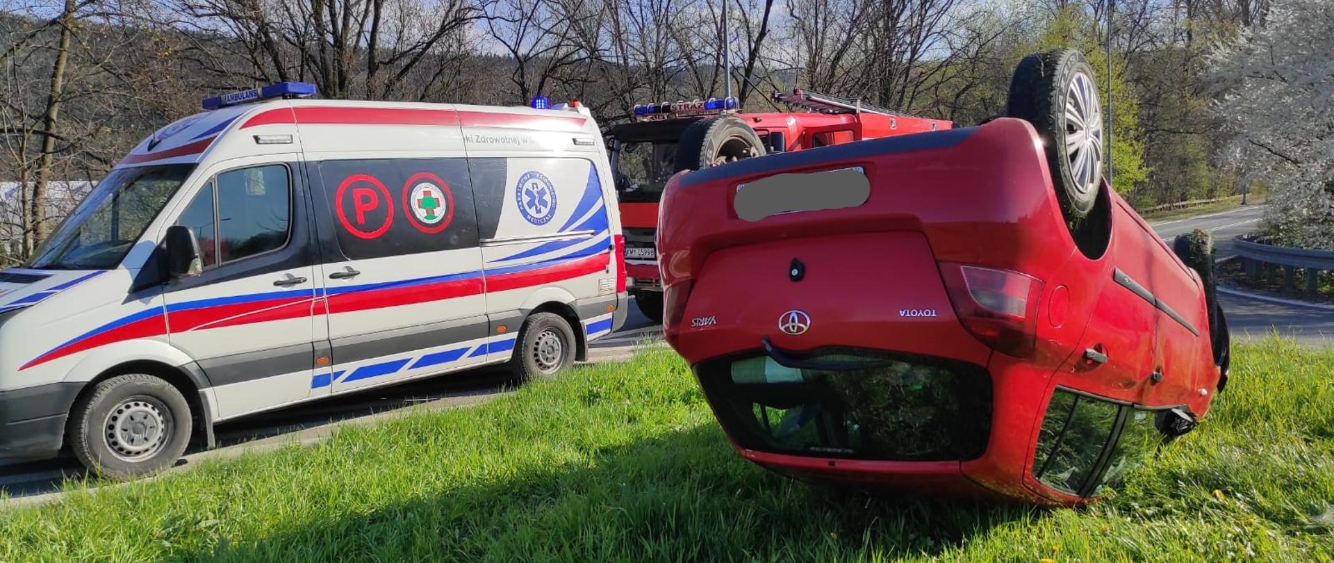 Na zdjęciu widzimy samochód osobowy w kolorze czerwonym leżący na dachu na trawniku przy zjeździe z drogi krajowej nr 7 w Myślenicach. Po lewej stronie zdjęcia widzimy stojącą karetkę pogotowia ratunkowego oraz stojący za nią samochód strażacki . W tle widoczne zarośla, drzewa rosnące wzdłuż drogi.