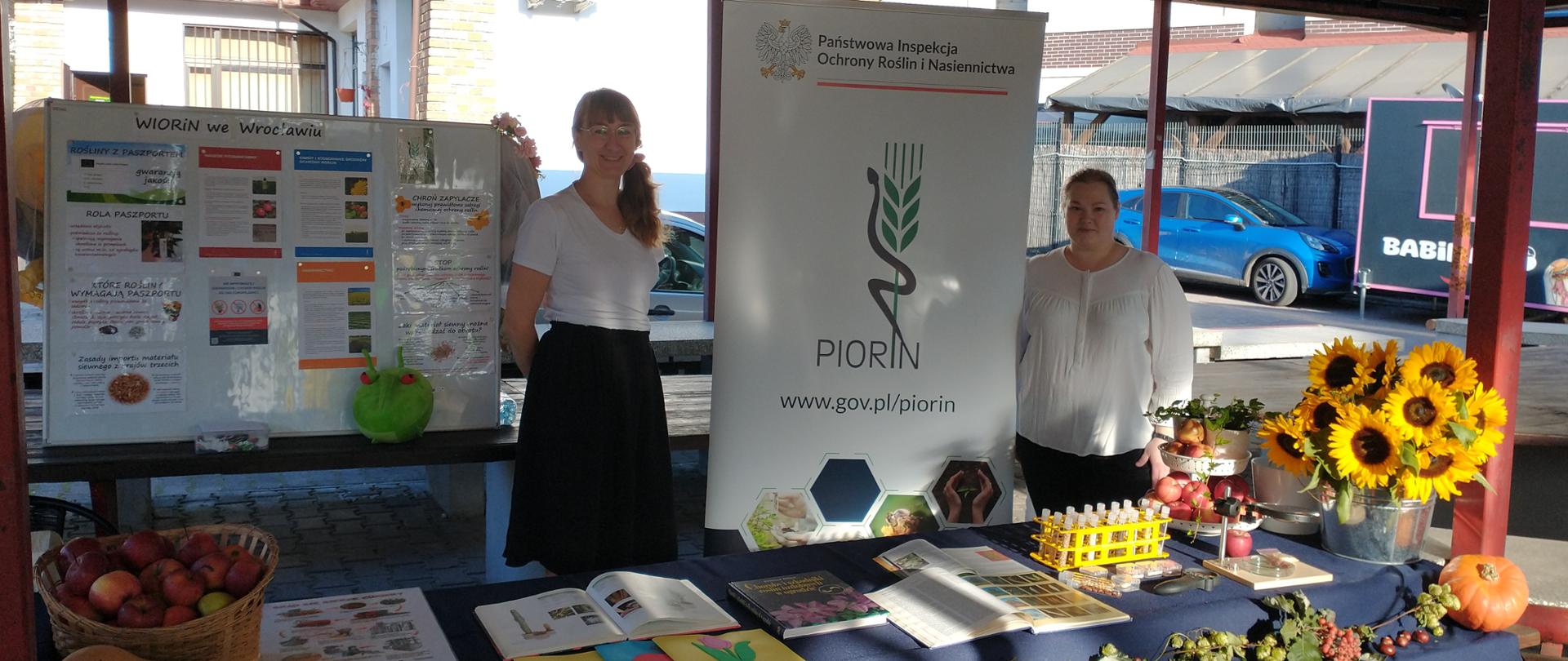 Dwie Panie Inspektor stojące na stoisku edukacyjnym. Na stole kolorowe materiały informacyjne, jesienne dekoracje w postaci, słoneczników, kosza z jabłkami, w tle baner i tablica informacyjna.