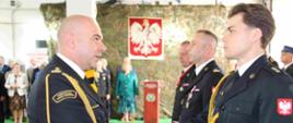 Zdjęcie w budynku. Na pierwszym planie dwóch funkcjonariuszy PSP w mundurach galowych, jeden z nich składa gratulacje drugiemu. W tle inni funkcjonariusze PSP i osoby cywilne. Na ścianie wisi siatka maskująca z zawieszonym Godłem Polski.