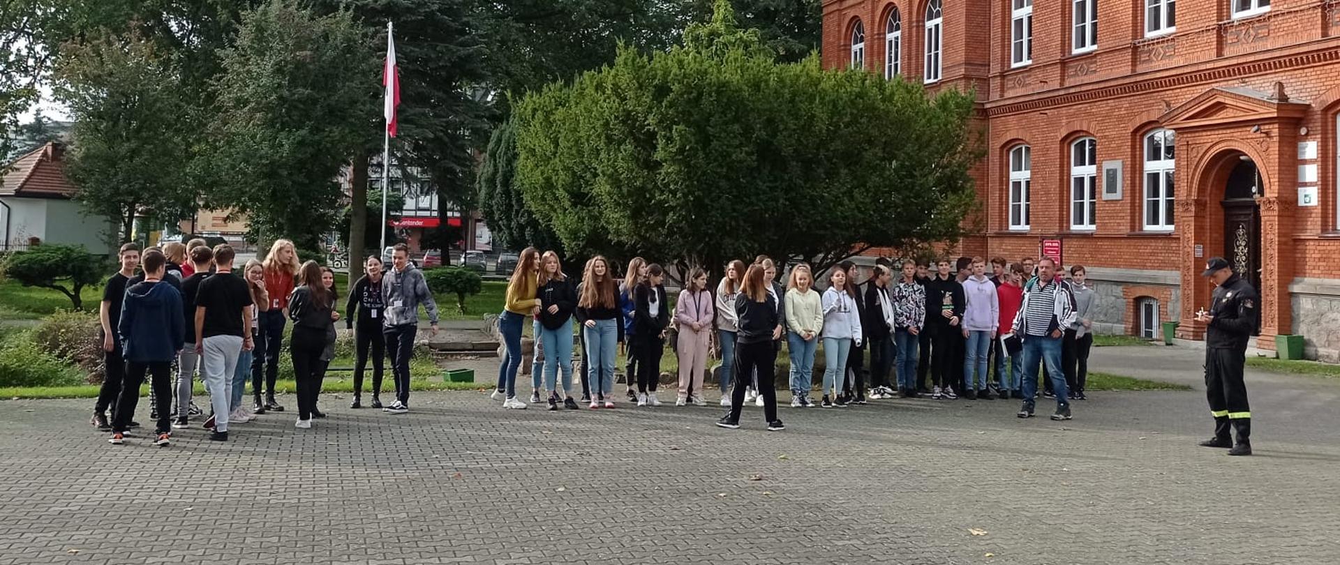 Zdjęcie przedstawia plac przed budynkiem z czerwonej cegły - Zespołem Szkół imienia Władysława Broniewskiego w Świdwinie na którym zgromadziła się młodzież. Strażak bacznie obserwuję realizację ewakuacji.