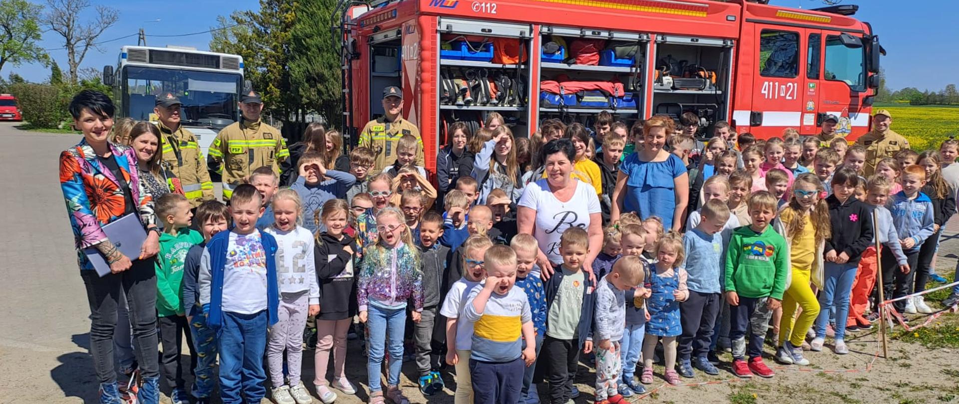 Na zdjęciu duża grupa dzieci i nauczycielek kolorowo ubrana. W tle samochód strażacki, obok strażacy.