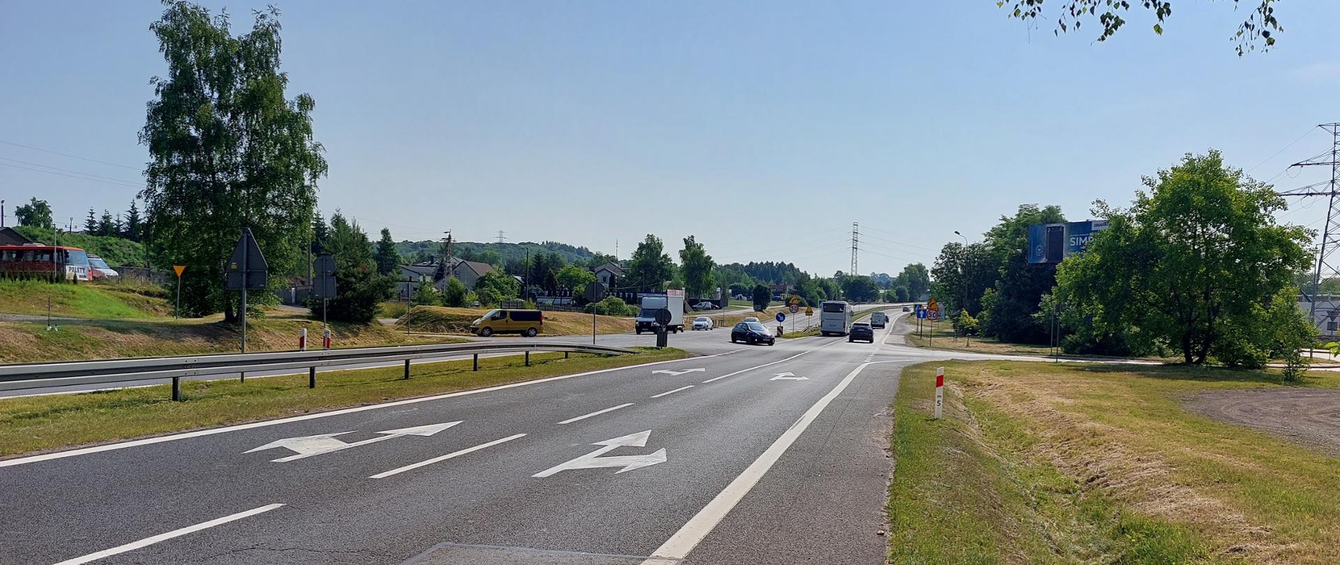 zdjęcie drogi krajowej nr 94 w Olkuszu. Widoczne dwie jezdnie po dwa pasy ruchu w obu kierunkach z oznakowaniem poziomym i pionowym. Jezdnie oddzielone są pasem zieleni i barierami ochronnymi. 