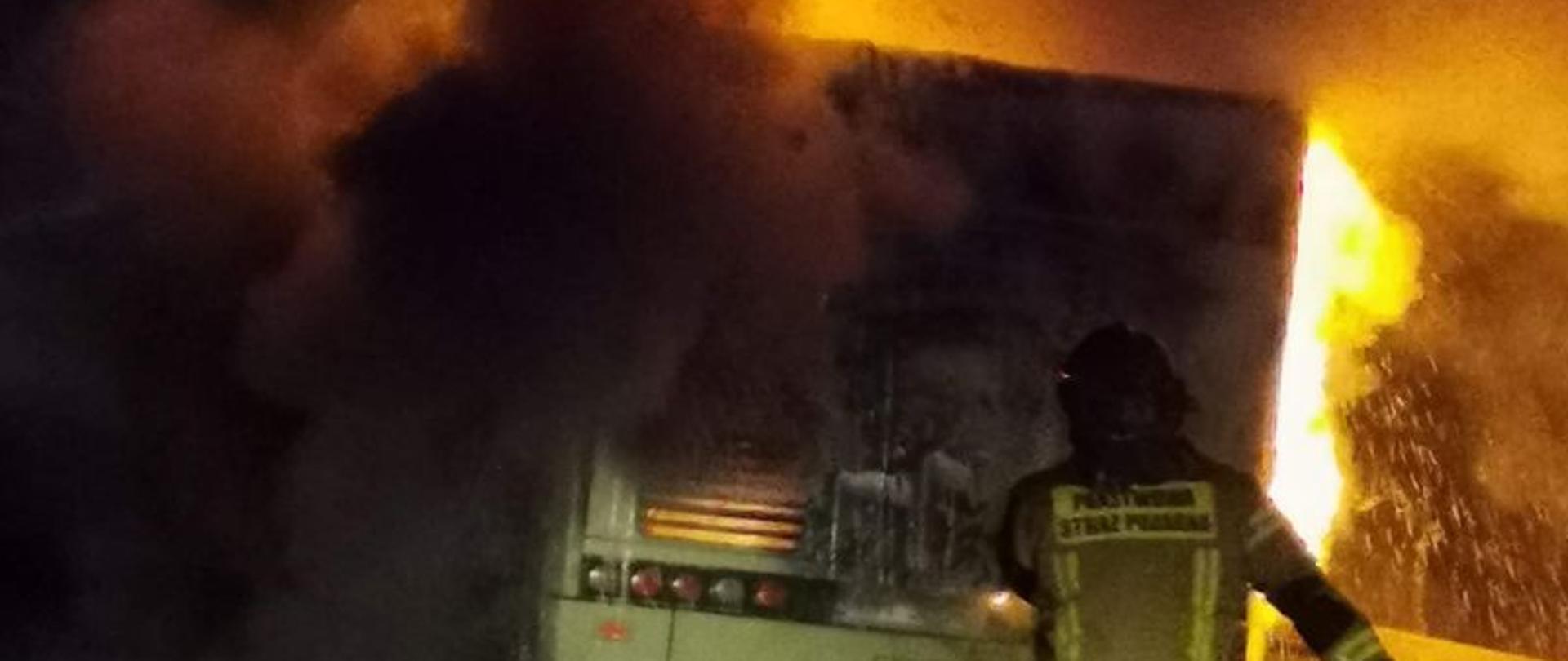 Pożar autobusu w fazie rozwiniętej. Widoczna duża ilość dymu i płomienie, na pierwszym planie strażak koordynujący działania.