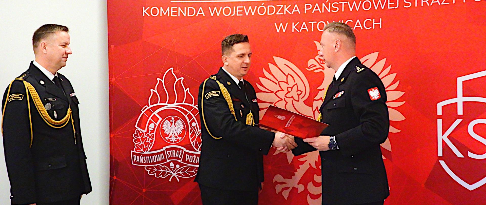 Trzej funkcjonariusze Państwowej Straży Pożarnej w galowych mundurach stoją przed czerwonym tłem z logo Komendy Wojewódzkiej PSP w Katowicach. Dwóch z nich podaje sobie ręce podczas wręczenia dokumentu, trzeci stoi obok, obserwując uroczystość.