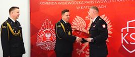 Trzej funkcjonariusze Państwowej Straży Pożarnej w galowych mundurach stoją przed czerwonym tłem z logo Komendy Wojewódzkiej PSP w Katowicach. Dwóch z nich podaje sobie ręce podczas wręczenia dokumentu, trzeci stoi obok, obserwując uroczystość.