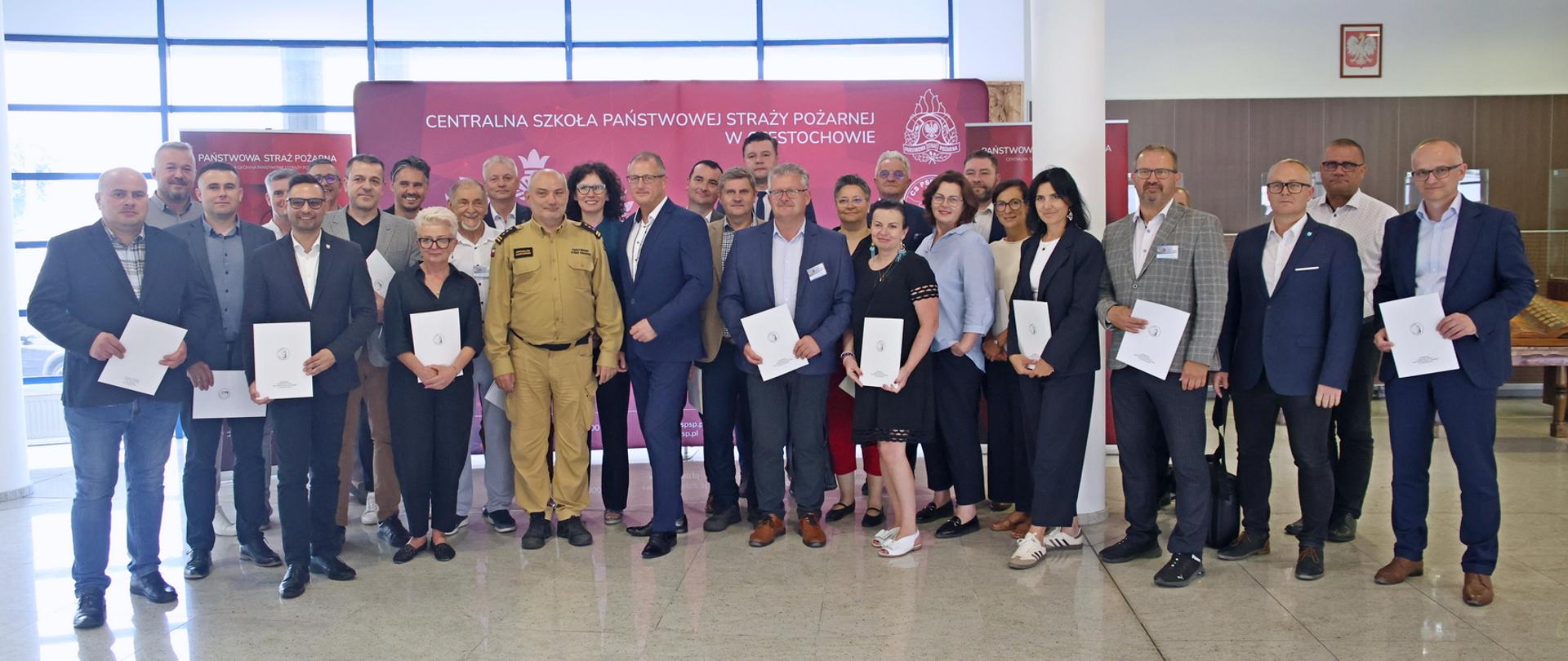 Pamiątkowe zdjęcie grupowe uczestników szkolenia z zakresu ochrony ludności i obrony cywilnej dla osób pełniących funkcję wójtów, burmistrzów i prezydentów miast (28 osób) na tle banneru Centralnej Szkoły PSP, w towarzystwie Komendanta Centralnej Szkoły PSP