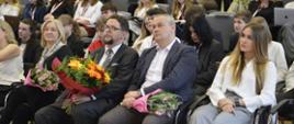 Wiceminister Szeptycki siedzi w otoczeniu kobiety i mężczyzny - wszyscy trzymają na kolanach bukiety kwiatów.