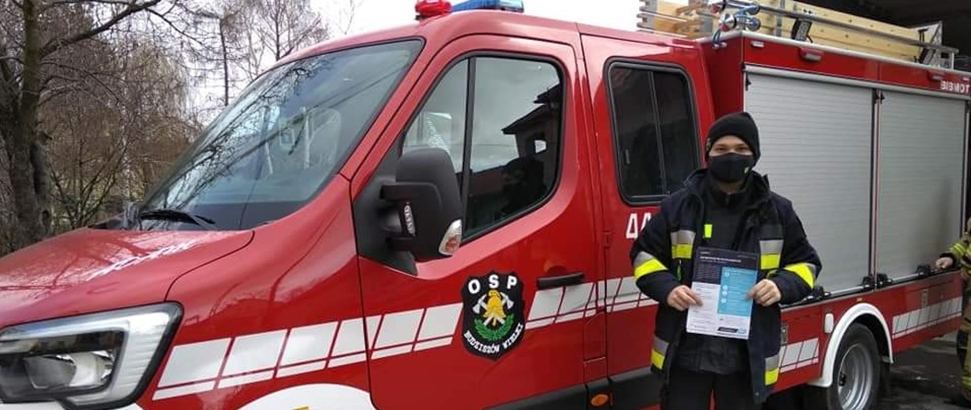 Obraz przedstawia strażaka prezentującego ulotkę informacyjną dotyczącą szczepienie przeciwko koronawirusowi. Strażak na tle samochodu pożarniczego.