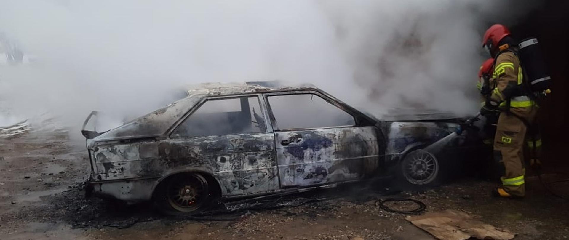 Na podwórku przy garażu 2 strażaków gasi palący się samochód osobowy. Samochód jest cały spalony. 