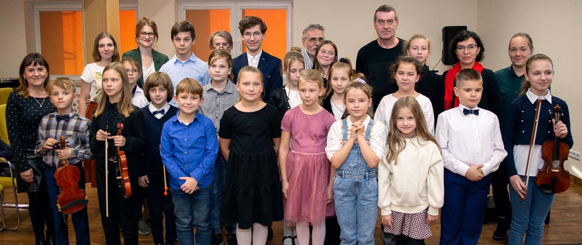 Na zdjęciu, w pomieszczeniu stoi grupa kilkunastu osób, dzieci i dorosłych, wszyscy patrzą do przodu, niektórzy z nich w rękach trzymają skrzypce.