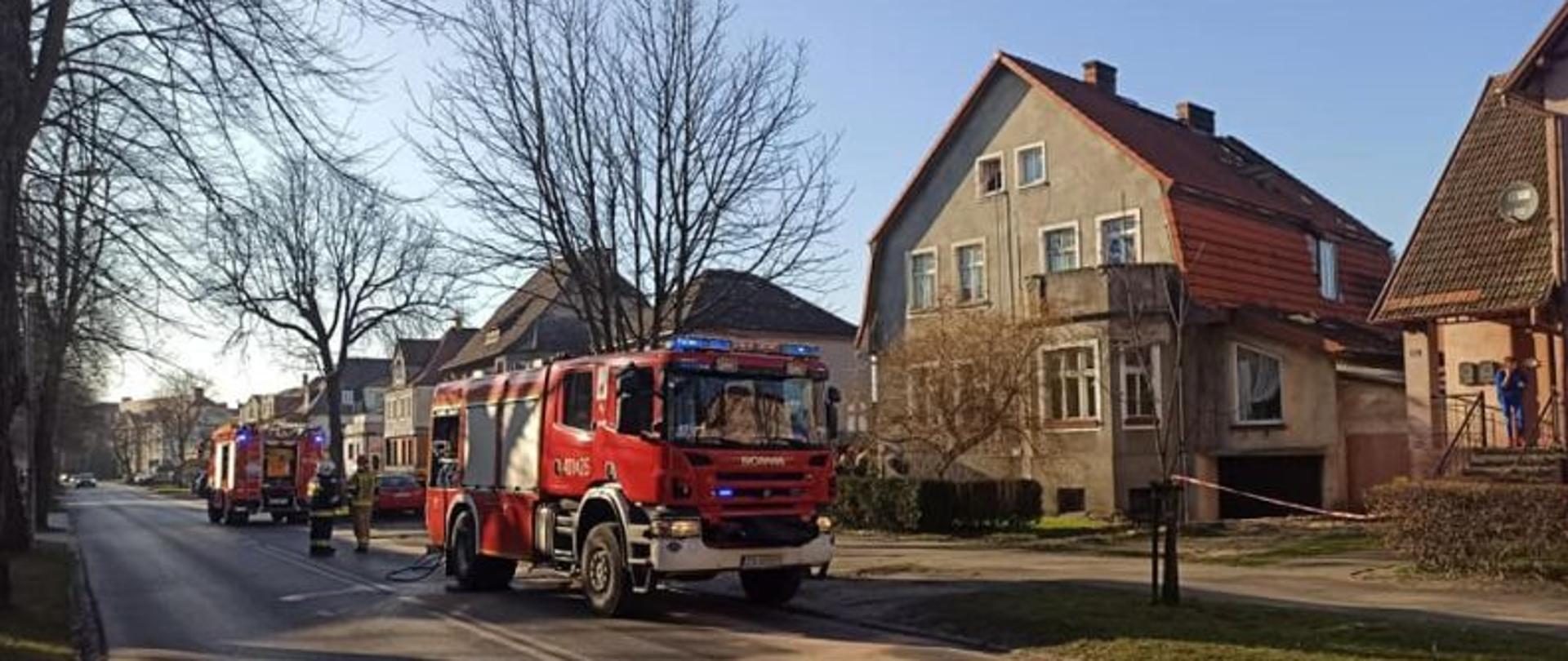 Zdjęcie wykonane w dzień. Na pierwszym planie stojące wzdłuż ulicy auta gaśnicze. W tle budynek objęty pożarem. Zdjęcie wykonane po ugaszeniu pożaru.