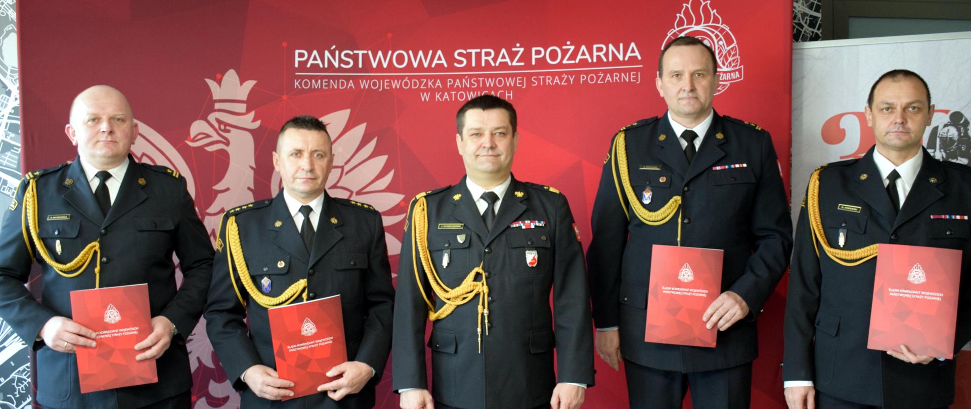 Na tle czerwonej ścianki z wizerunkiem orła i napisem u góry Państwowa Straż Pożarna Komenda Wojewódzka PSP w Katowicach stoją w mundurach wyjściowych: w środku śląski komendant wojewódzki PSP po jego prawej i lewej stronie czterech powołanych zastępców komendantów powiatowych i miejskich PSP.