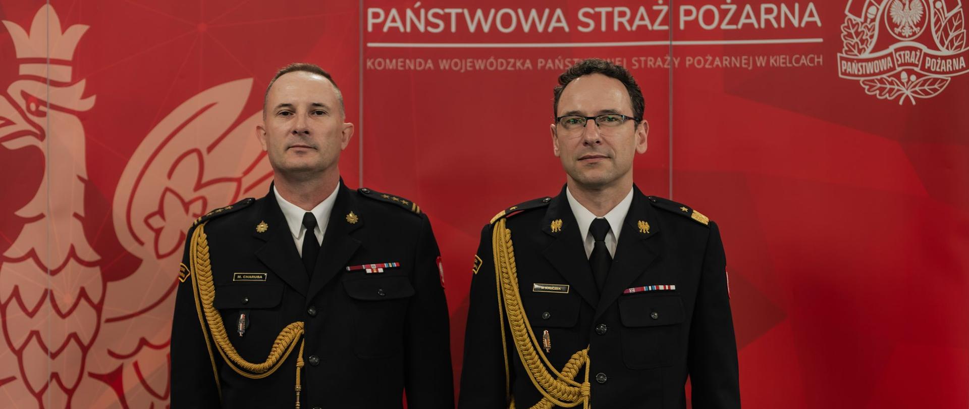Dwóch funkcjonariuszy PSP pozuje na tle ścianki z godłem i logotypem Państwowej Straży Pożarnej. Obaj w pełnym galowym umundurowaniu z żółtymi sznurami.