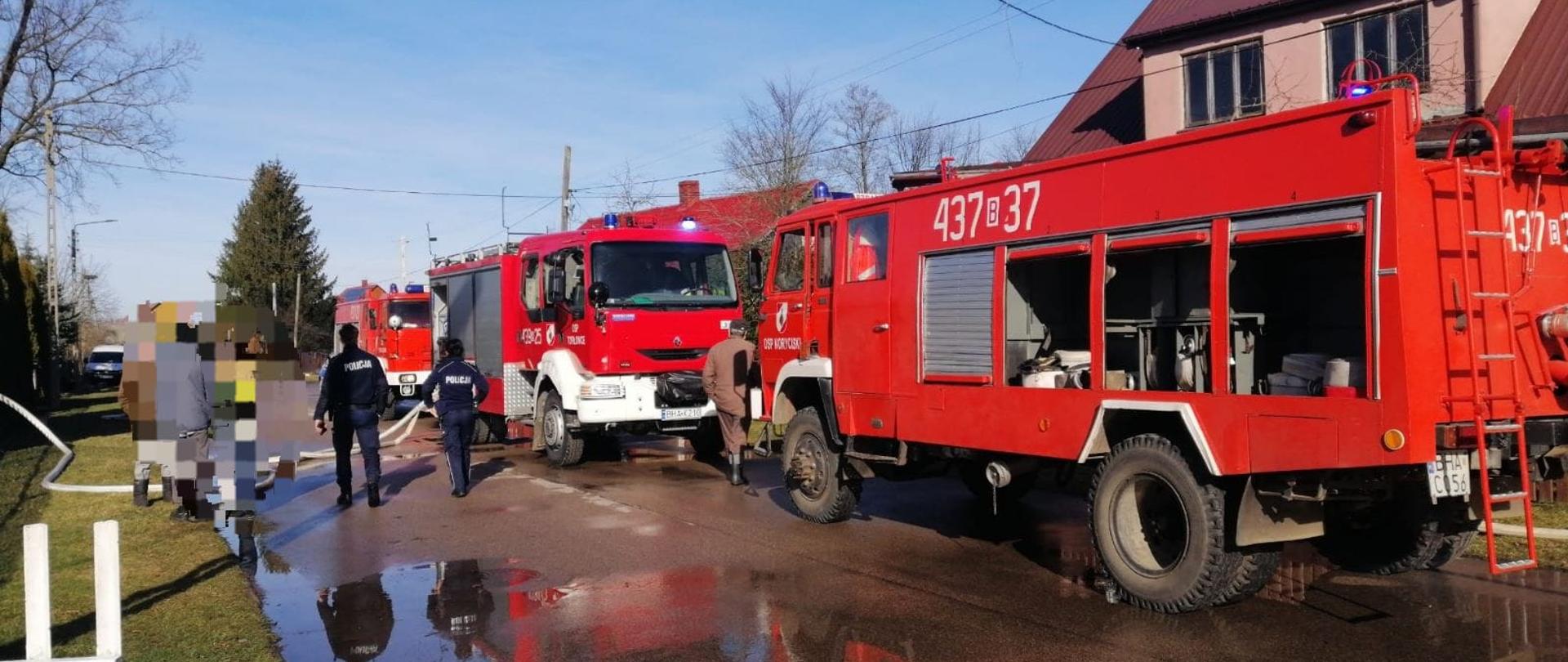 Na ulicy stoi kilka strażackich wozów bojowych. Obok stoją policjanci.