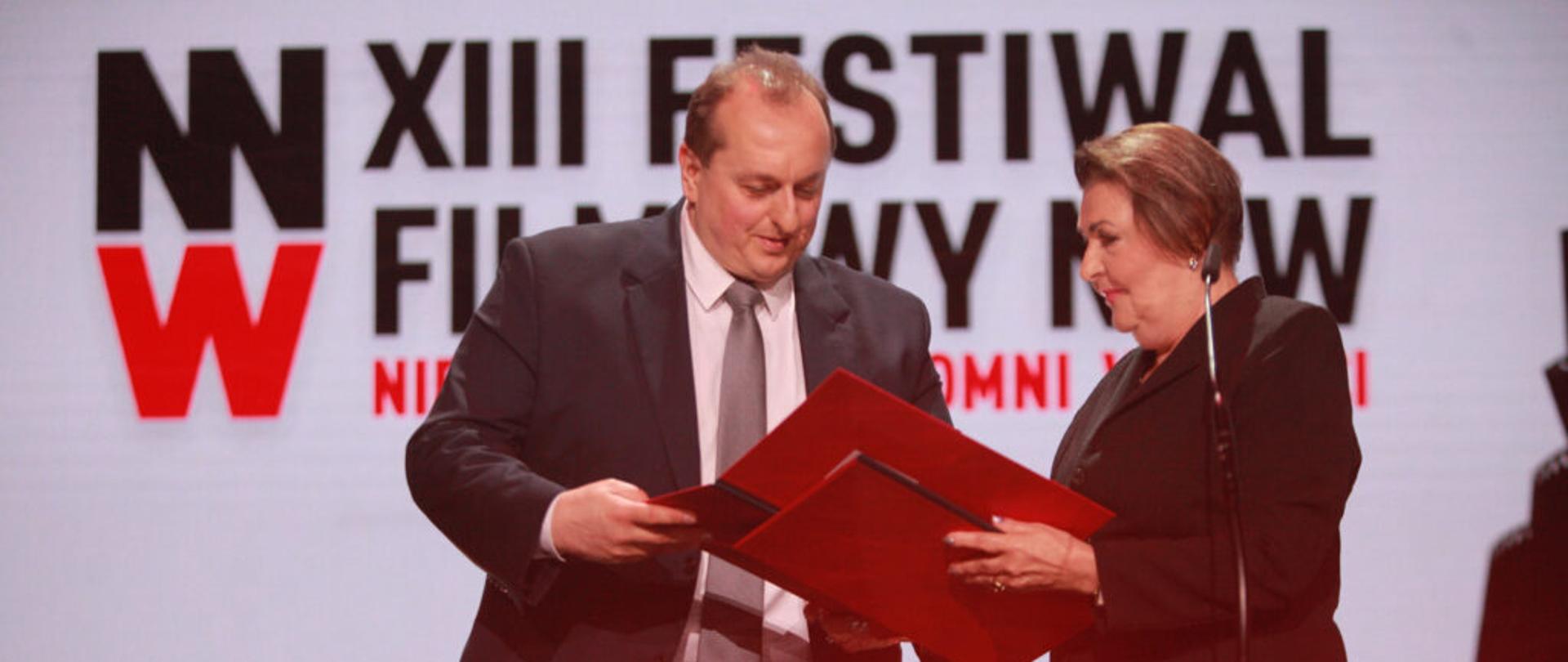 Zdjęcie ilustracyjne w poziomie. Członek KRRiT Elżbieta Więcławska-Sauk stoi na scenie, wręcza dyplom laureatowi. W tle ekran z czerwonym napisem na białym tle - "XIII Festiwal Filmowy NNW"