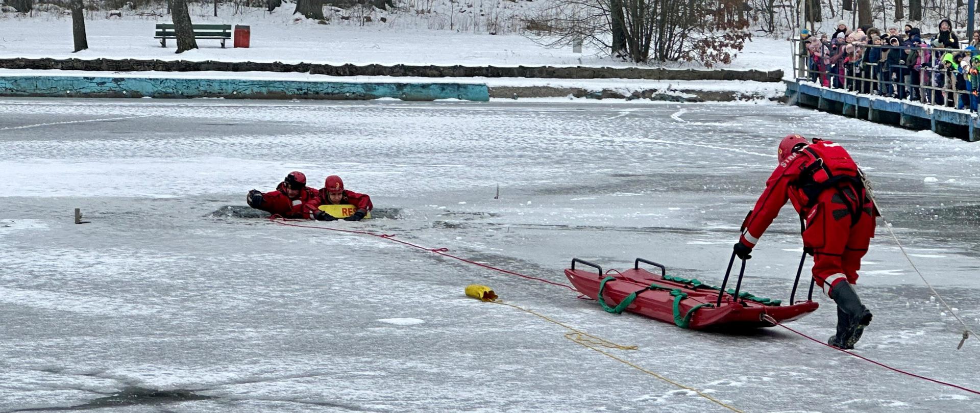 Zdjęcie dokumentuje kluczowe fazy pokazu ratownictwa wodno-lodowego, przeprowadzonego przez strażaków PSP w ramach kampanii „Bezpieczne Ferie 2026”.
Faza 1: Dotarcie do poszkodowanego i zabezpieczenie
Na szerszym ujęciu widać moment, w którym ratownicy znajdują się bezpośrednio przy przeręblu:
Bezpośrednia pomoc: Dwóch ratowników w czerwonych kombinezonach znajduje się w wodzie lub przy jej krawędzi, zabezpieczając osobę poszkodowaną za pomocą żółtej bojki ratunkowej z napisem „RESCUE”.
Przygotowanie transportu: Trzeci ratownik na lodzie manewruje saniami lodowymi, ustawiając je tak, aby umożliwić sprawne podjęcie poszkodowanego z wody na pokład sań.
System liny: Przez taflę lodu przebiega długa, czerwona lina, która łączy zespół ratowniczy z brzegiem, zapewniając stałą kontrolę nad ich pozycją.
Faza 2: Ewakuacja na saniach lodowych
Drugie zdjęcie prezentuje techniki transportu po kruchym lodzie:
Wykorzystanie sań: Ratownicy umieścili „poszkodowanego” na czerwonych saniach lodowych. Jeden z nich leży na saniach wraz z ewakuowaną osobą, stabilizując jej pozycję.
Praca ratownika: Ratownik stojący przy uchwytach sań kontroluje ich bieg, podczas gdy zespół na brzegu (niewidoczny na tym zbliżeniu) przyciąga sprzęt za pomocą liny.
Zabezpieczenie: Wszyscy strażacy operujący na lodzie są wyposażeni w suche skafandry, kaski oraz buty zapewniające przyczepność.
Znaczenie edukacyjne
Oba zdjęcia ukazują, jak duża grupa widzów obserwuje akcję z bezpiecznej odległości na pomoście. Pokazy te wizualizują trudności związane z ratowaniem osób pod którymi załamał się lód, podkreślając, że bez specjalistycznych narzędzi, takich jak sanie lodowe czy kombinezony wypornościowe, taka akcja jest ekstremalnie ryzykowna.