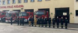 Strażacy z JRG stoją w szeregu na tle pojazdów pożarniczych i budynku jednostki. 
