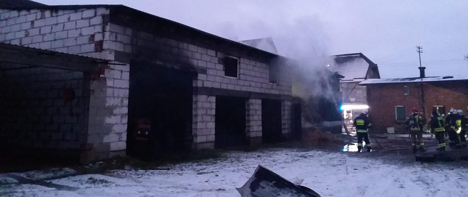 Strażacy za pomocą prądów wodnych gaszą pomieszczenia gospodarcze, z których wydobywa się dym.