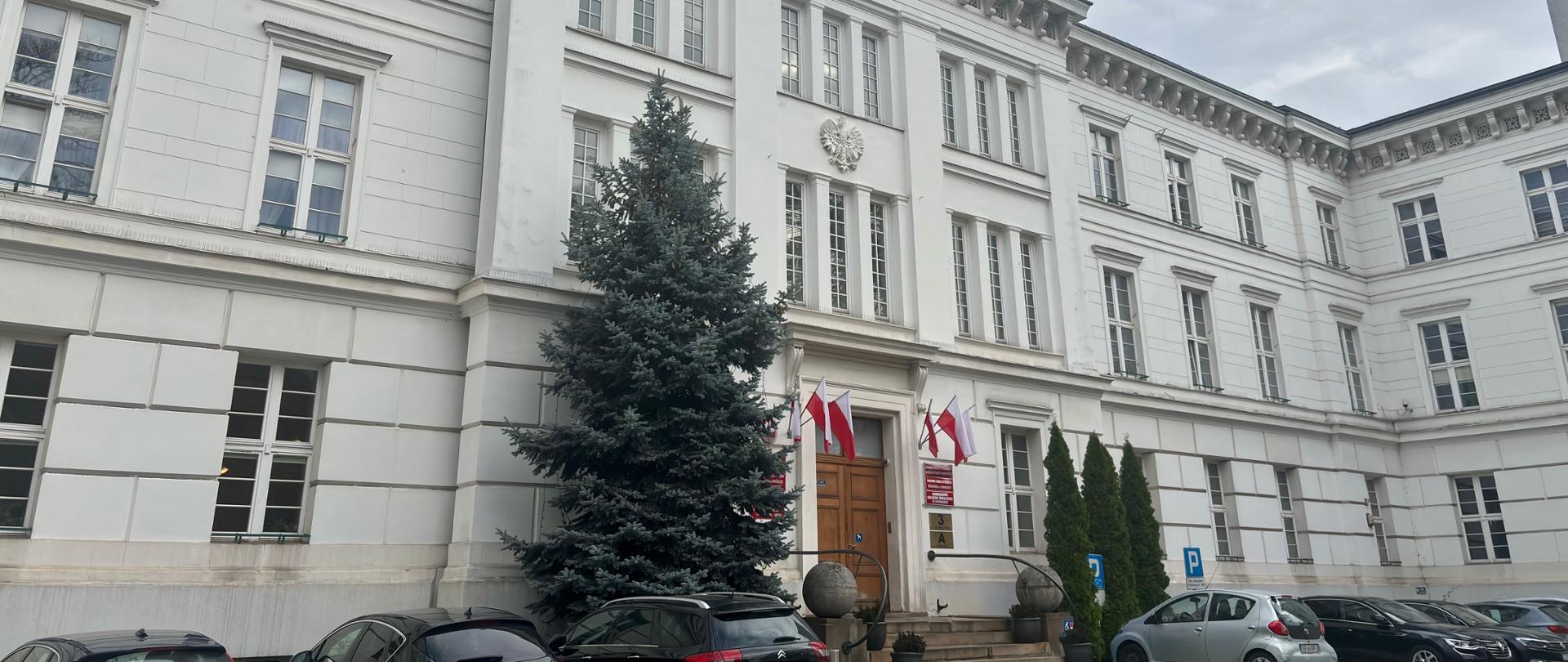 gmach Kujawsko-Pomorskiego Urzędu Wojewódzkiego, przy głównych dużych drewnianych drzwiach, po lewej i po prawej stronie flagi Polski, po lewo od drzwi wysoki zielony świerk.