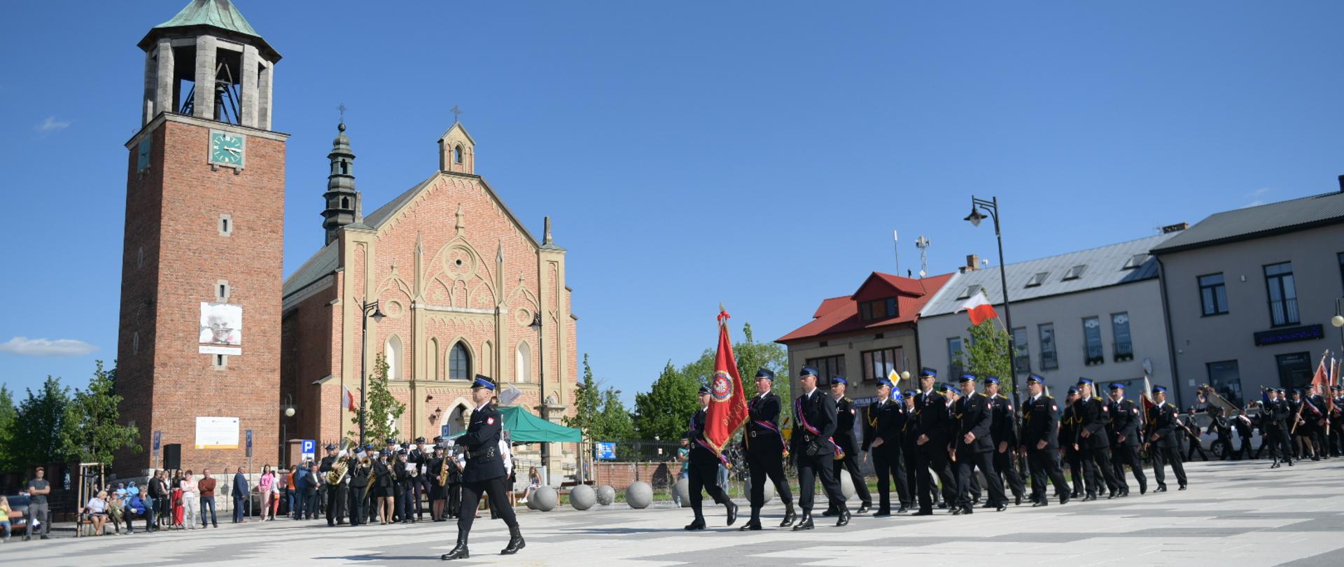 Strażacy maszerują po płycie rynku w Proszowicach, w tle kościół