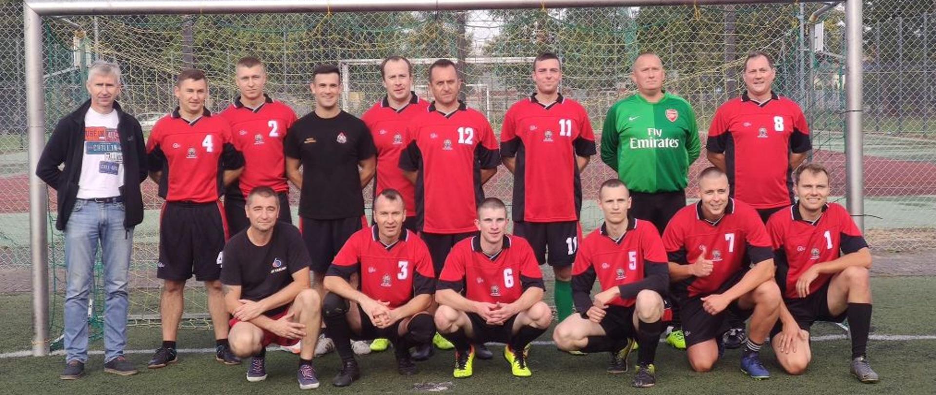 Zdjęcie grupowe drużyny KP PSP Białogard. W tle bramka. Strażacy w strojach sportowych czerwono czarnych.