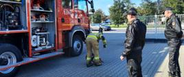 Na zdjęciu widać wóz strażacki oraz strażaka uruchamiającego pilarkę przed komisją przeglądową 