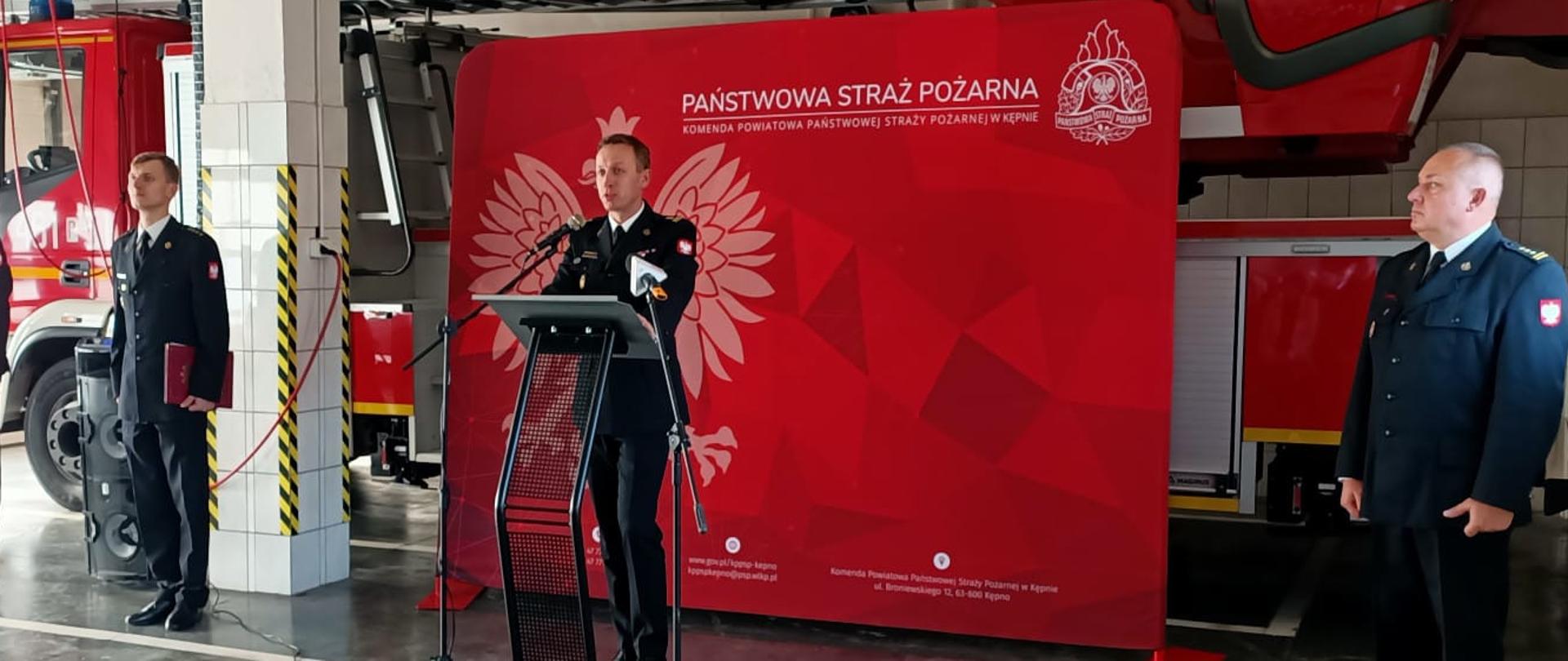 w garażu strażak stoi przed mównicą, w tle pojazd pożarniczy, obok strazaka stoją inni strażacy