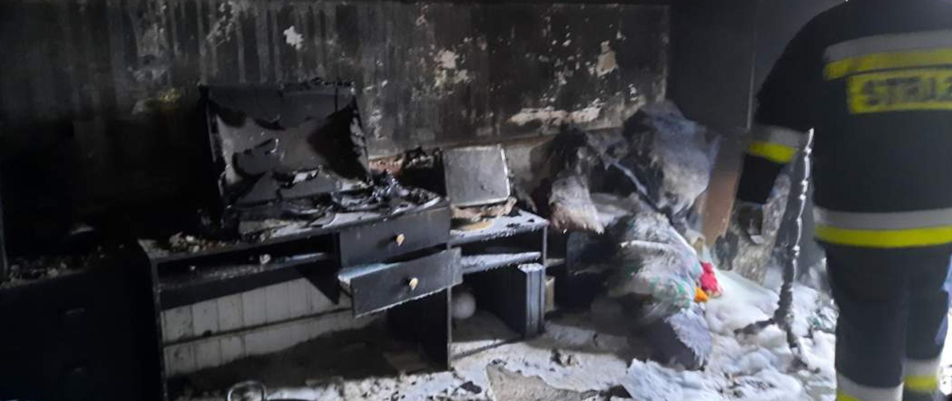 Zdjęcie przedstawia spalone pomieszczenie, pokój po ugaszeniu pożaru. W pomieszczeniu znajdują się zniszczone, częściowo spalone meble, okopcone ściany i sufit.