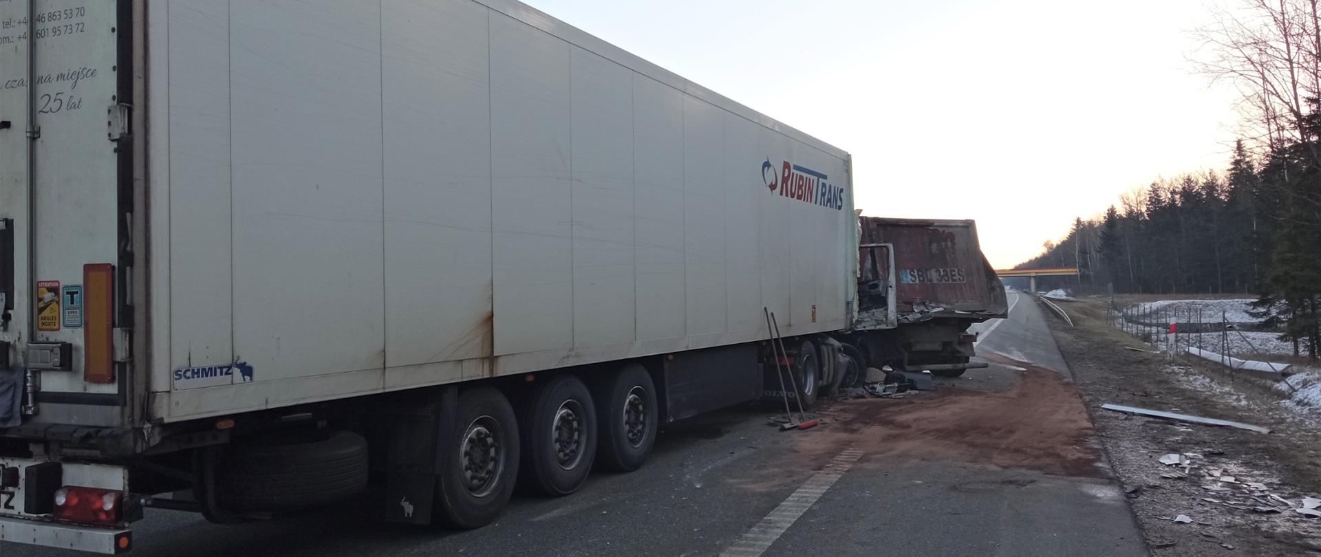 Na zdjęciu widzimy samochody ciężarowe, które uległy zderzeniu. Na pierwszym planie ogromna skrzynia izotermy. Na dalszym planie plama płynów eksploatacyjnych posypana sorbentem i fragment drugiego pojazdu. W perspektywie droga S7