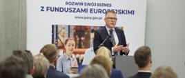 P.o. prezesa PARP Krzysztof Gdula stoi przy mównicy i przemawia do mikrofonu, za jego plecami baner informujący o rozwoju biznesu z Funduszami Europejskimi 