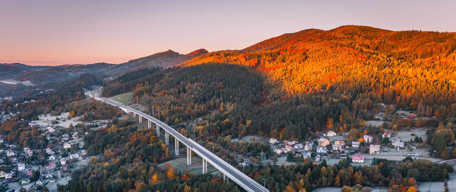 Widok z lotu ptaka na długi estakadę drogową biegnącą przez górską dolinę. Most przecina zalesione wzgórza oraz niewielką miejscowość, a jesienne słońce oświetla lasy i zbocza intensywnymi odcieniami pomarańczu i złota. Wokół widać domy, drogi oraz fragmenty pól pokrytych porannym szronem.