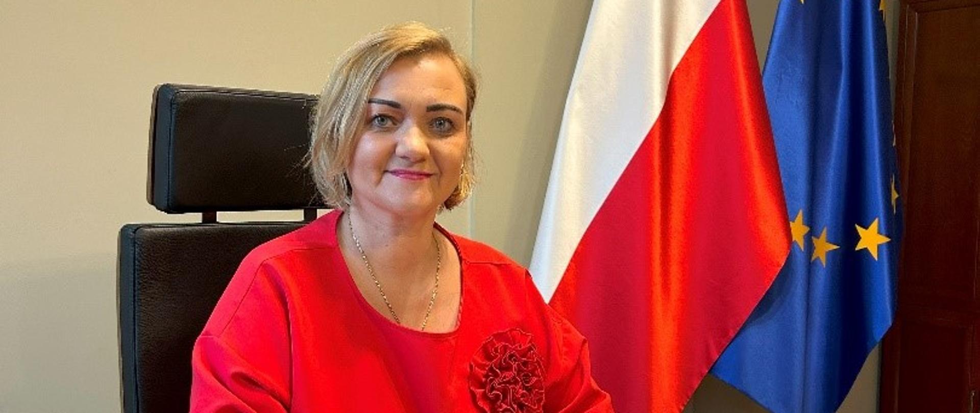 Zdjęcie przedstawia kobietę siedzącą przy biurku. W tle widoczne są flagi Polski i Unii Europejskiej