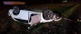 Na zdjęciu widać rozbity biały samochód, który leży na dachu w rowie. Ma otwarte przednie drzwi. Widać trawę oraz elementy karoserii samochodu na poboczu. Jest ciemno.