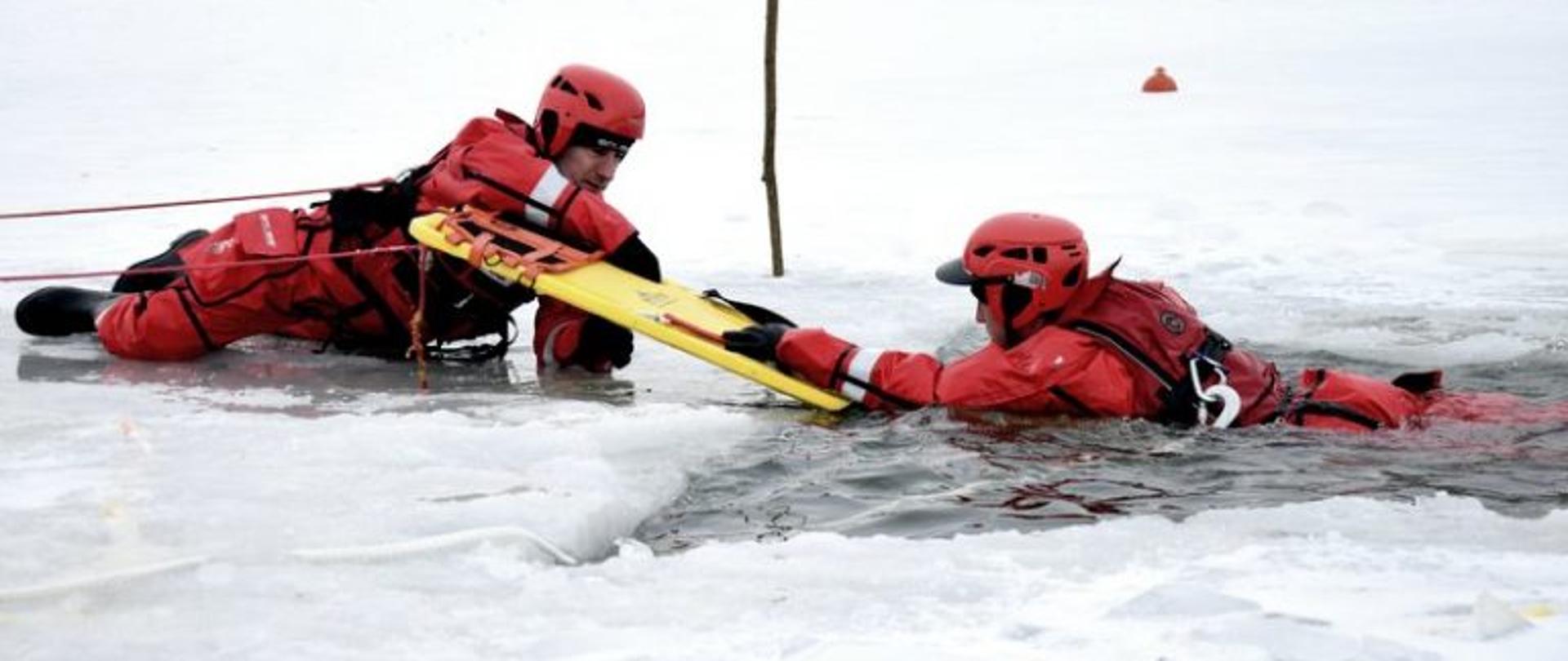 Zdjęcie przedstawia dwóch strażaków ubranych w skafandry suche oraz kaski. Jeden ze strażaków znajduje się w wodzie, natomiast drugi leży w wodzie i za pomocą deski ratowniczej próbuje wyciągnąć go z wody. Tło stanowi śnieg